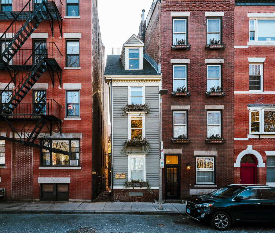 A narrow wooden house with pretty window boxes wedged tightly between two much larger red brick apartments buildings